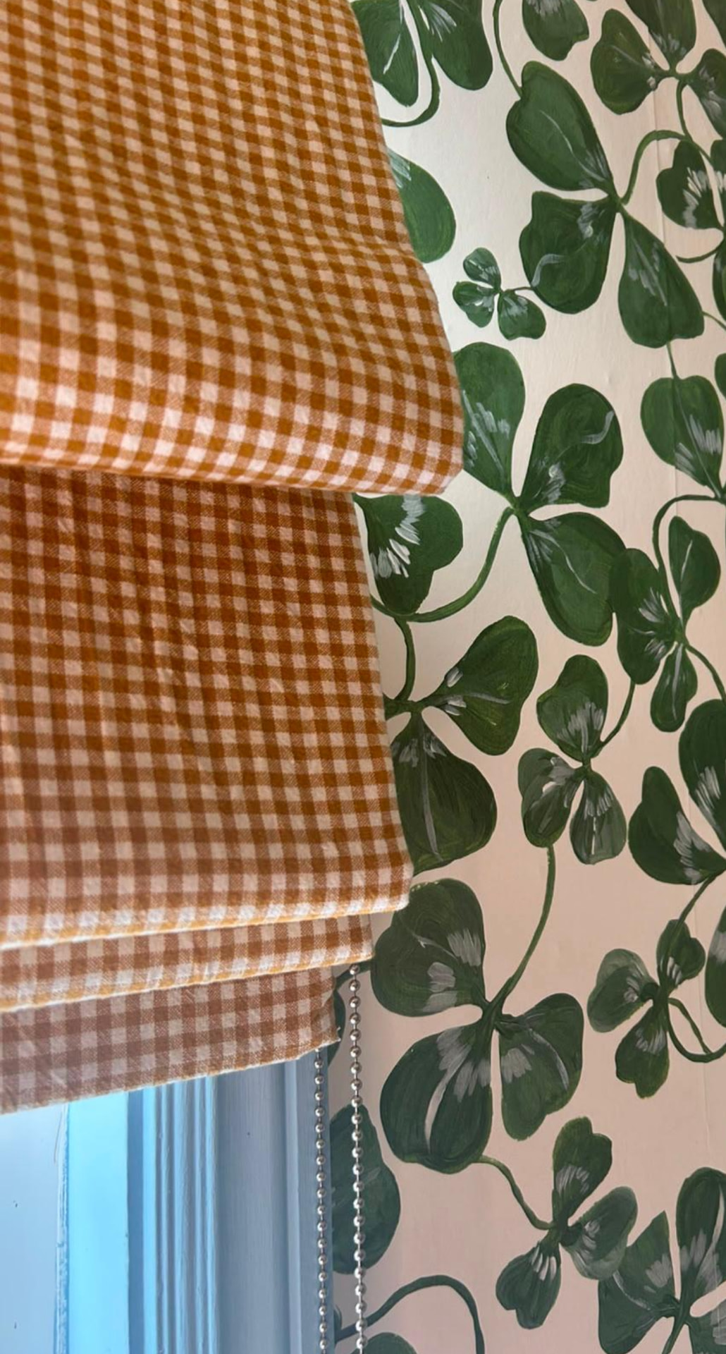 A Roman blind in terracotta gingham hanging at a window beside green clover wallpaper